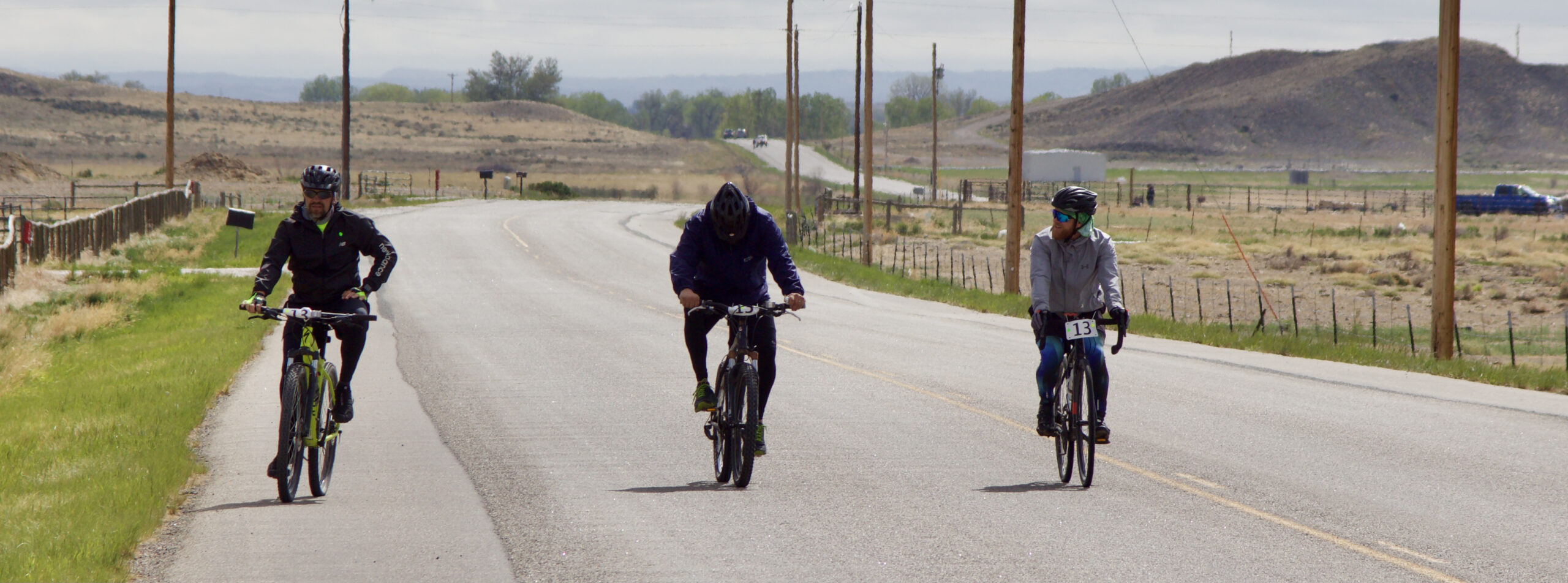 Three people biking on a street outside of town with mountains in the background.