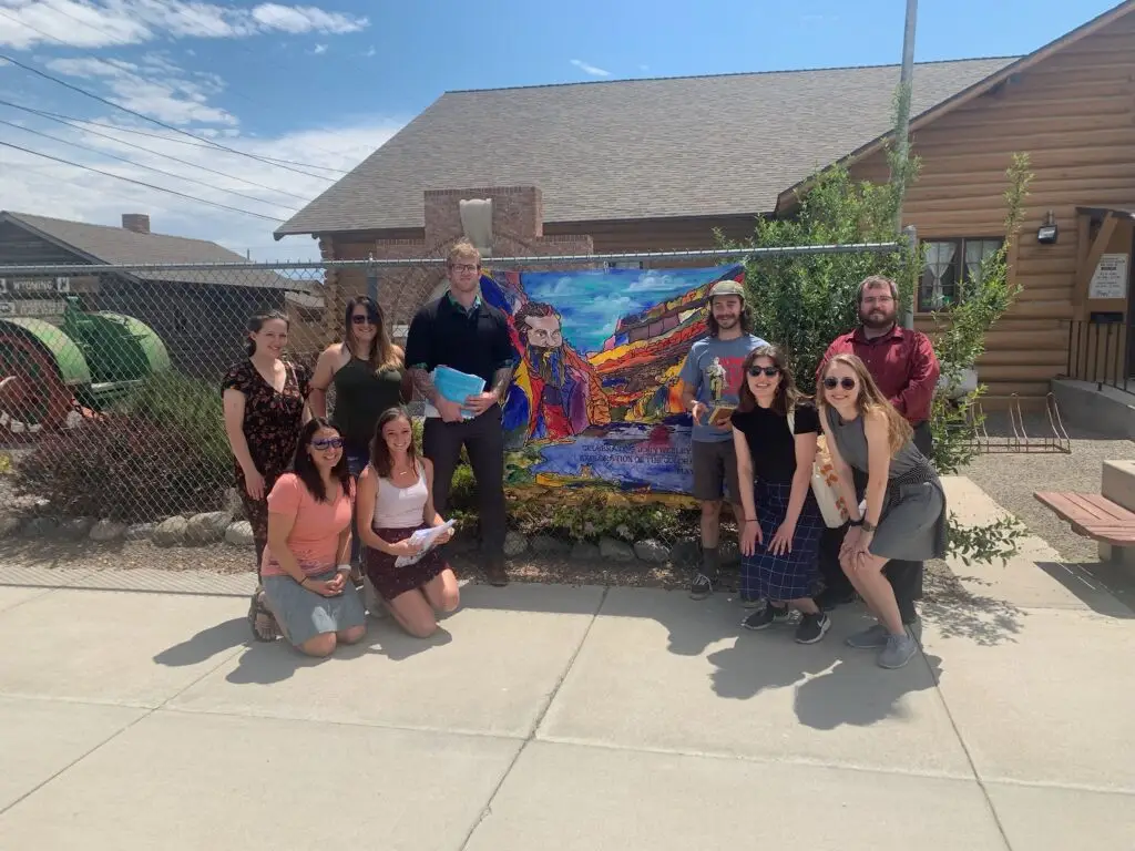 AmeriCorps VISTA Members posing in front a log cabin style building with a mural of John Wesley Powell hung on a fence