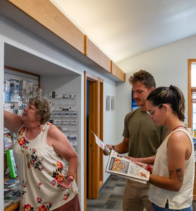 An older woman grabbing reading material from a wall for a man and a younger woman who are reading from a magazine catalogue featuring businesses, events, and other info in Powell, Wyoming